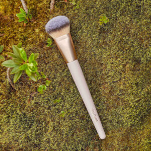 Der Make up Pinsel DOLO PINSEL GRUNDIERUNG 01 mit beigem Griff und goldener Zwinge liegt auf einer moosigen Oberfläche, umgeben von kleinen grünen Pflanzen.