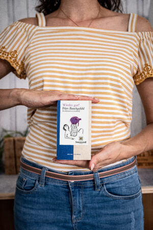 Eine Person im gestreiften Hemd hält ein Buch mit dem Titel „SONNENTOR WIEDER GUT! FÜRS BAUCHGEFÜHL TEE“ mit Illustration und Text auf dem Cover vor einem hölzernen Hintergrund.