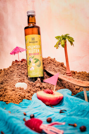 Eine Flasche SONNENTOR HOLUNDERBLÜTEN SIRUP steht mit einer Spielzeugpalme, Sonnenschirmen, einer halbierten Nektarine und Dekorationen mit Strandmotiven vor einem pastellfarbenen Hintergrund auf Sand.