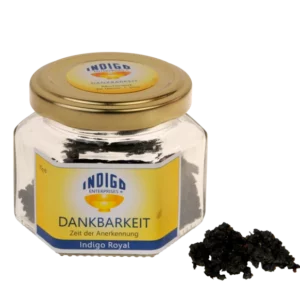 Ein sechseckiges Gefäß mit goldenem Deckel und der Aufschrift „Indigo Enterprises DANKBARKEIT – Zeit der Anerkennung“ enthält eine dunkle Substanz, von der ein kleiner Teil außerhalb des Gefäßes zu sehen ist.