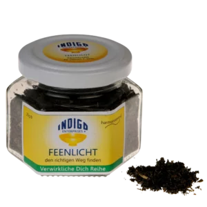 Sechseckiges Gefäß mit der Aufschrift „FEENLICHT – den richtigen Weg finden“ von Indigo Enterprises, gefüllt mit dunklen Teeblättern. Daneben liegen einige Blätter verstreut.