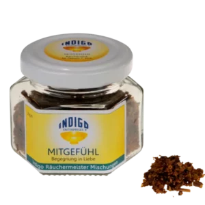 Ein kleines Glasgefäß mit der Aufschrift „MITGEFÜHL – Begegnung in Liebe“ enthält eine getrocknete Kräutermischung, daneben steht ein kleiner Haufen der Mischung.