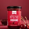 Ein Glas XUCKER FRUCHT KIRSCH 10091 Kirschfruchtaufstrich mit Xylit mit der Aufschrift "80% weniger Zucker" steht auf einer roten Fläche neben frischen Kirschen.