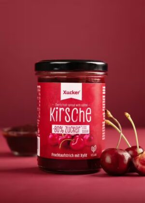 Ein Glas XUCKER FRUCHT KIRSCH 10091 Kirschfruchtaufstrich mit Xylit mit der Aufschrift "80% weniger Zucker" steht auf einer roten Fläche neben frischen Kirschen.