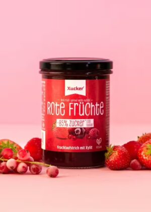 Ein Glas XUCKER FRUCHT ROT FR 10092 Fruchtaufstrich mit 85% weniger Zucker steht auf einer rosafarbenen Fläche, umgeben von Erdbeeren, Johannisbeeren und Himbeeren vor einem rosafarbenen Hintergrund.