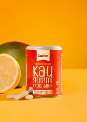 Eine Dose XUCKER XYLIT KAUG FRUC 10174 Kaugummi steht vor einer Zitronenspalte und einer Mango auf einem leuchtend gelben Hintergrund.