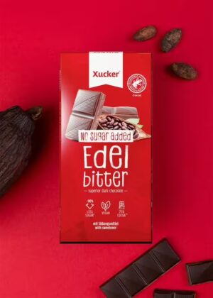 Eine rote Schachtel XUCKER VEG EDELB XYLIT 10149, eine hochwertige Zartbitterschokolade ohne Zuckerzusatz, die neben Kakaobohnen und Schokoladenstücken auf rotem Hintergrund abgebildet ist.