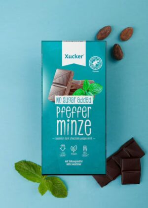 Schachtel mit XUCKER VEG EDELB PFEF 10137 Pfefferminzschokolade ohne Zuckerzusatz, umgeben von Schokoladenstücken, Minzblättern und Kakaobohnen auf blauem Hintergrund.