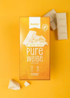 Auf gelbem Hintergrund ist eine Schachtel XUCKER WEISS SCHOKO 10138 abgebildet, daneben liegen weiße Schokoladenstücke.
