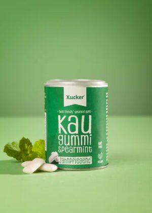 Ein grüner Kanister mit der Aufschrift "XUCKER XYLIT KAUGUMMI 10170" ist neben weißen Kaugummistücken und Minzblättern auf grünem Hintergrund zu sehen.