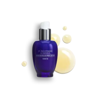 Eine blaue Flasche IMMORTELLE PRECIEUSE SERUM von LOccitane mit einer weißen Pumpe ist von gelben Flüssigkeitströpfchen umgeben.