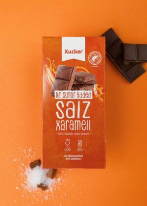 Eine orangefarbene Schachtel mit XUCKER XYL SCHOKO SALZ 10133 Milchschokolade steht auf einem orangefarbenen Hintergrund, daneben liegen Schokoladenstücke und ein kleiner Haufen Salz.