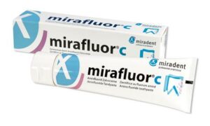 Vor der Verpackung steht eine Tube ZP.MIRADENT MIRAFLUOR CR C-Zahnpasta mit blau-weißem Aufdruck und dem Miradent-Logo.