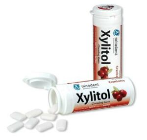 Zwei Packungen MIRADENT XYLIT.GUM CRANBERRY, wobei eine Packung geöffnet ist und Kaugummistücke herausfallen.