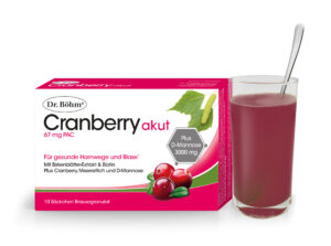 Eine Packung DR.BÖHM CRANBER.AKUT BRGRAN steht neben einem Cranberry-Getränk. Auf der Verpackung steht ein Text zu Cranberry und D-Mannose, ergänzt durch Cranberry-Bilder und grüne Blätter.